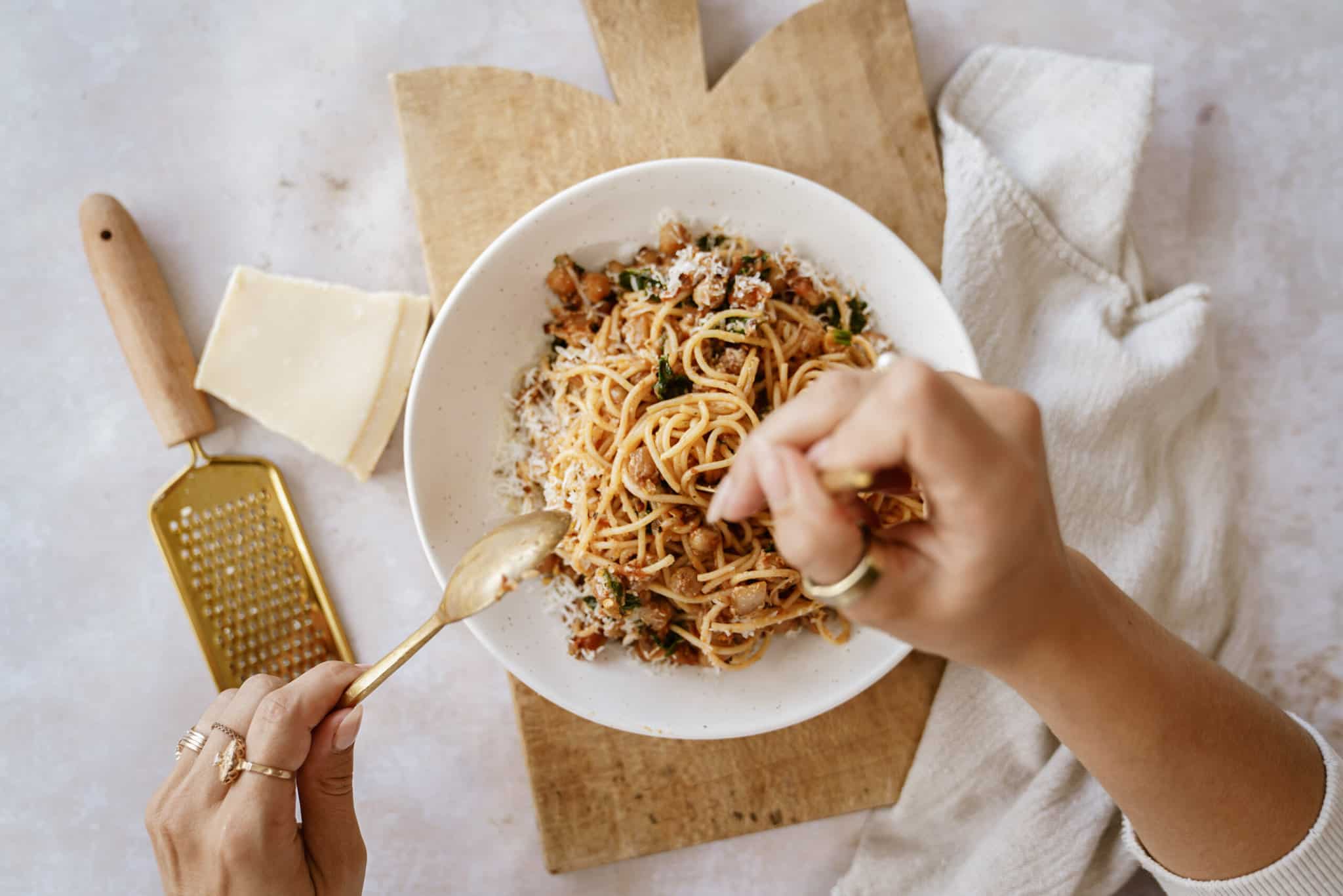 Hands using a gold fork to twirl chickpea pasta in a bowl. On the side is a block of parmesan and a hand cheese grater.