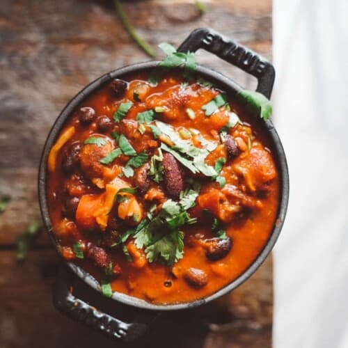 A pot of rich, red chili on a wooden surface, garnished with fresh cilantro. The chili contains beans and vegetables, adding texture and color. Bright natural light highlights the dish, with a sprig of cilantro nearby.