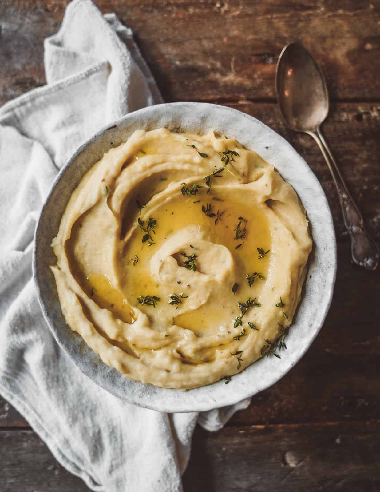 A bowl of creamy mashed potatoes garnished with fresh herbs, placed on a rustic wooden table. A folded white cloth and a tarnished silver spoon lie next to the bowl. The scene has a cozy, warm ambiance.