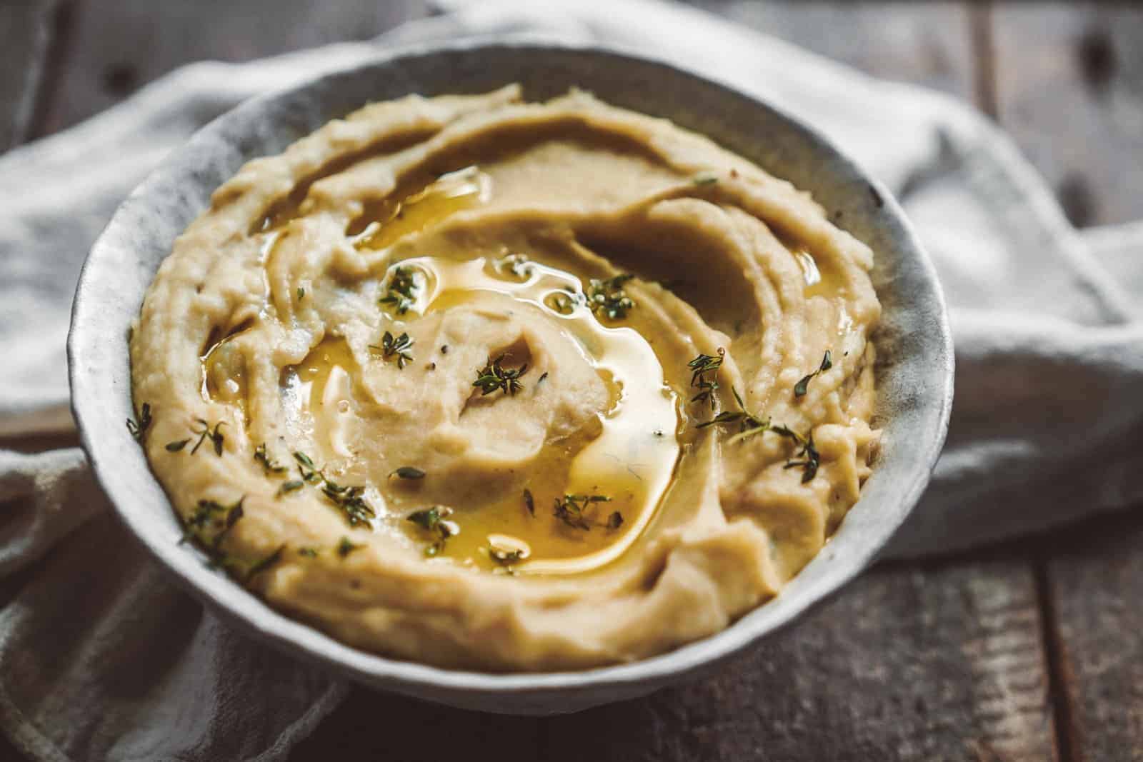 A bowl of creamy mashed potatoes topped with olive oil and garnished with fresh thyme. It is placed on a rustic wooden table with a white cloth underneath.