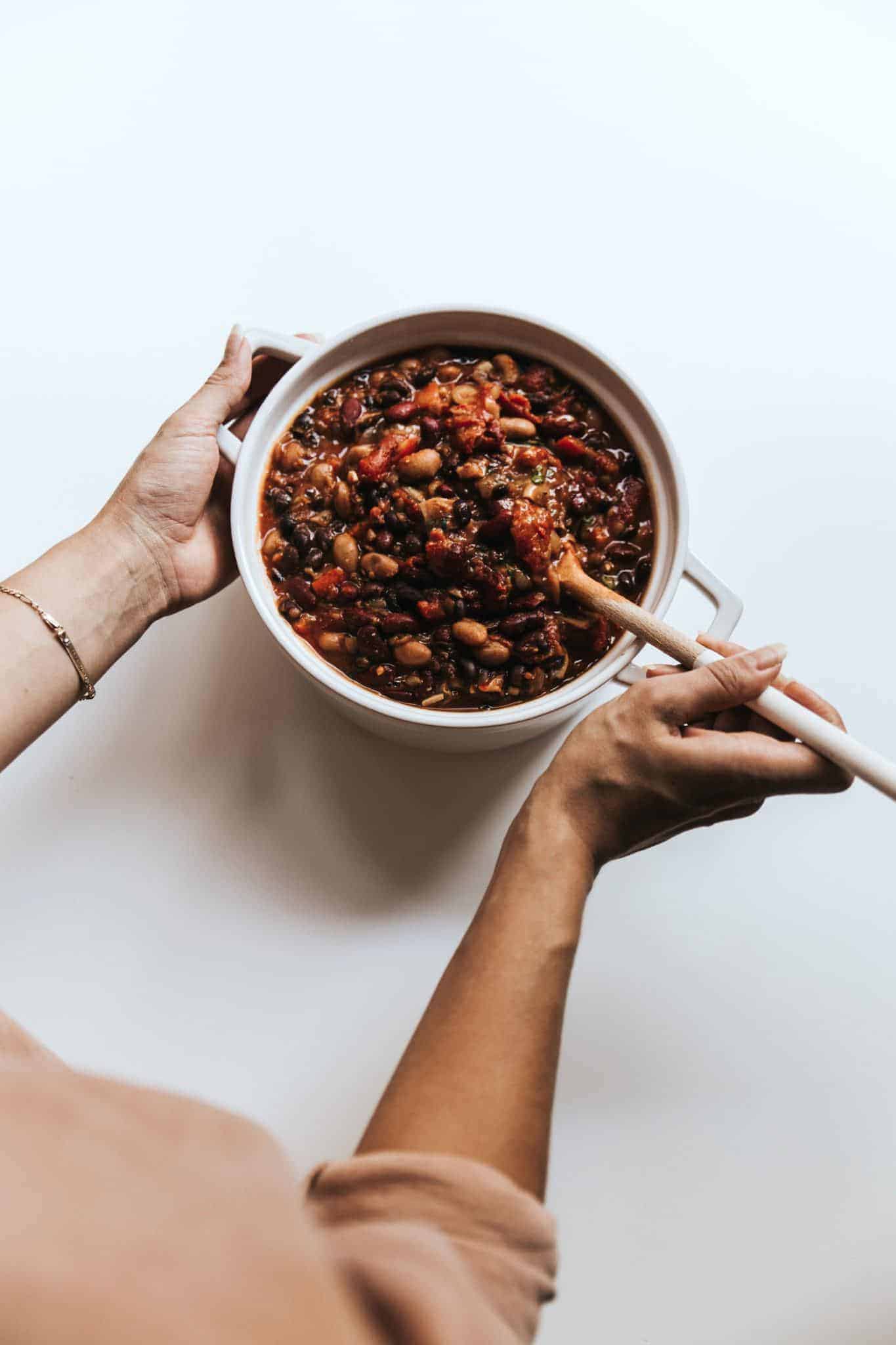 A hand stirs a pot of bean and lentil chili with a wood spoon.