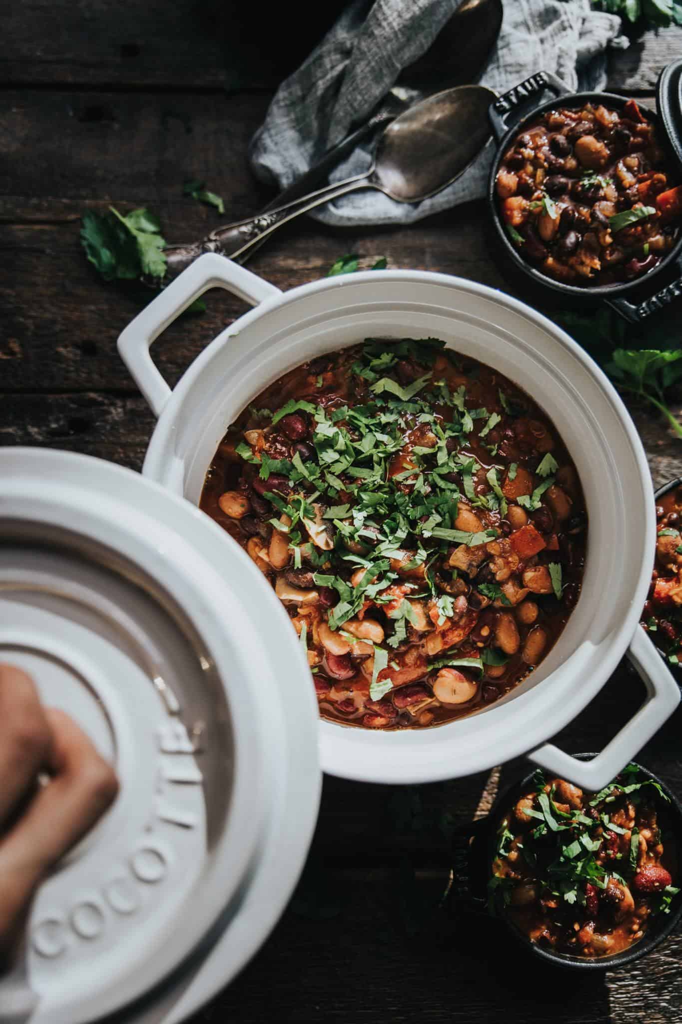 A hand open the lid of a white cast iron pot filled with lentil and bean chili, topped with colorful herbs.