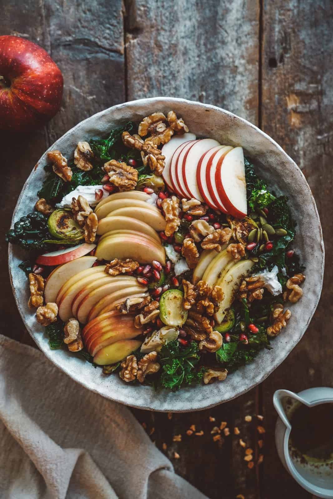 A rustic bowl filled with fresh salad, featuring sliced apples, kale, walnuts, Brussels sprouts, and scattered pomegranate seeds. Its set on a wooden table with a whole apple and a small pitcher nearby.