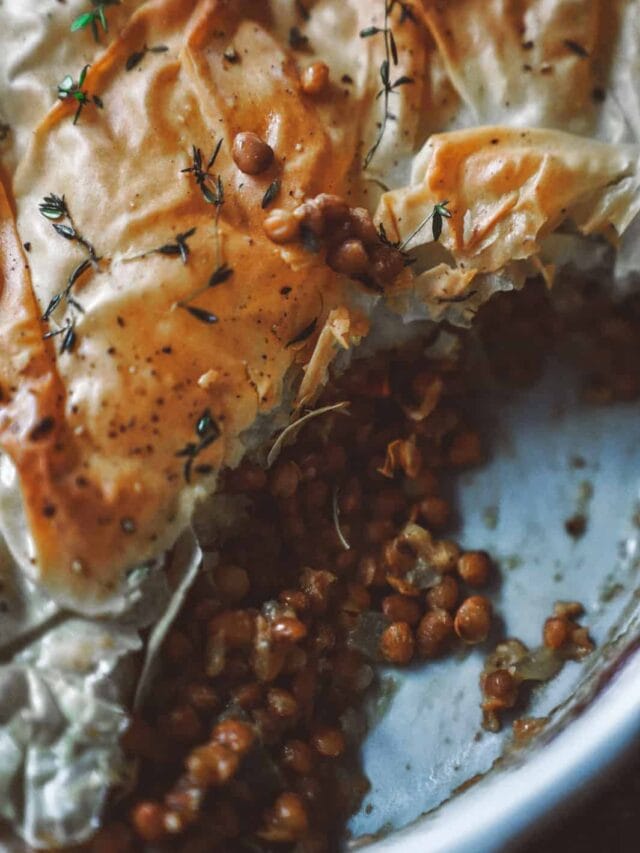 Close-up of a golden-brown pastry crust, partially broken to reveal a filling of cooked lentils. The dish is garnished with fresh thyme on top, providing a rustic and appetizing presentation in a ceramic dish.