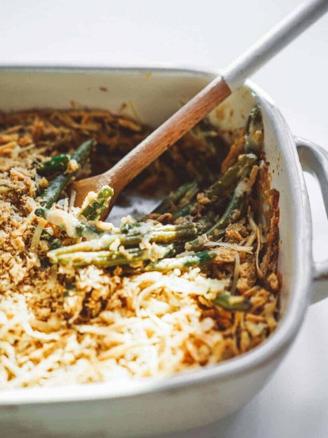 A creamy green bean casserole in a white baking dish, topped with melted cheese and crispy breadcrumbs. A wooden spoon is nestled in the dish, suggesting it has been served from.