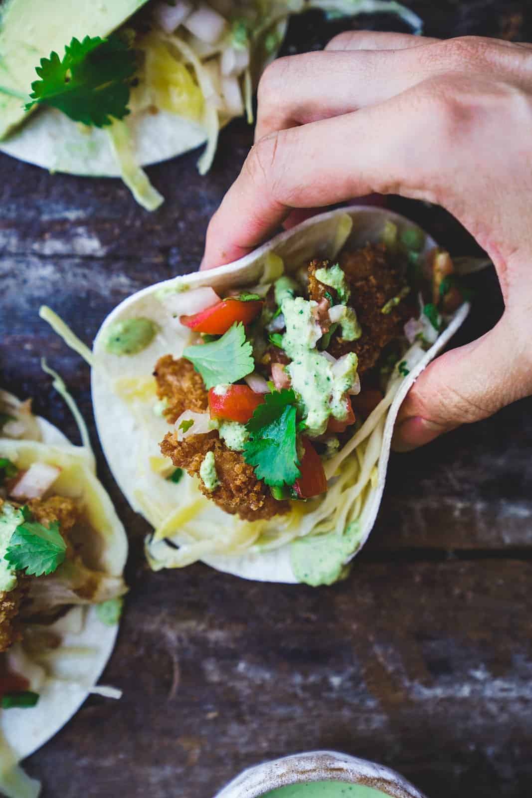 Hand holding a taco filled with golden-brown crispy cauliflower pieces, topped with shredded cabbage, fresh pico de gallo, and a drizzle of creamy cilantro sauce.