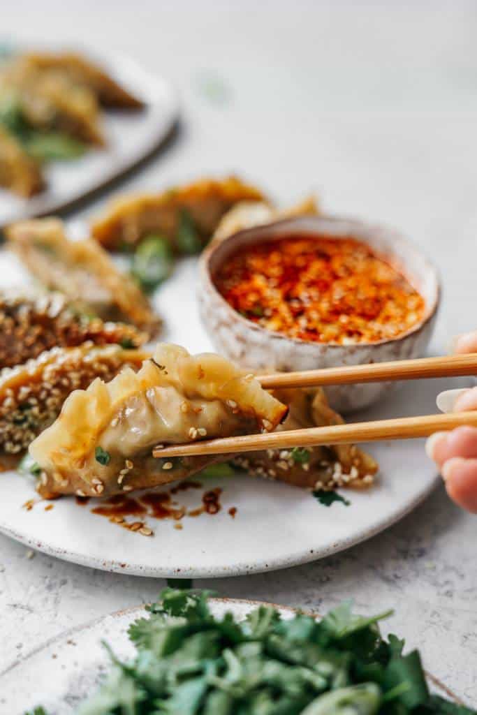 A hand using chopsticks to hold a dumpling over a white plate with more dumplings. A small bowl of chili oil dipping sauce is on the plate. Fresh cilantro is in the foreground on a grey surface.