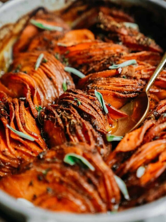 Close-up of a dish featuring thinly sliced, baked sweet potatoes with crispy edges, garnished with fresh herbs. A golden spoon rests among the potatoes, hinting at serving. The dish has a warm, inviting appearance.