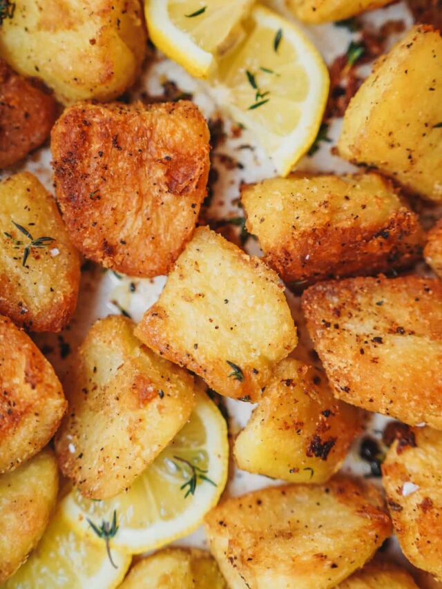 Close-up of crispy roasted potato chunks seasoned with herbs and spices, garnished with lemon wedges and small thyme leaves. The potatoes have a golden-brown crust, creating a rustic and appetizing appearance.