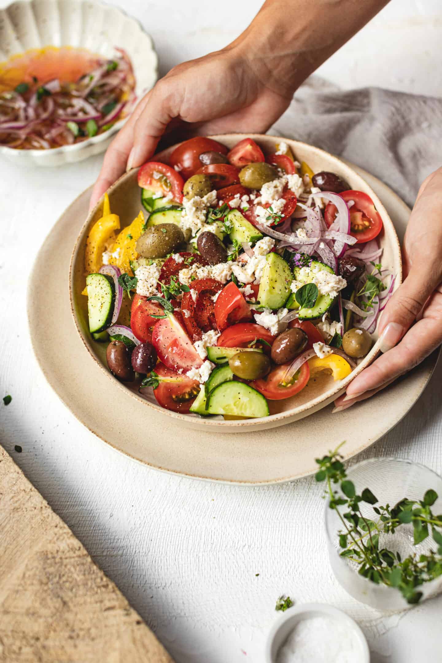 A vibrant Greek salad with tomatoes, cucumbers, olives, onions, and feta cheese sits in a bowl. Two hands are holding the dish on a neutral tabletop, surrounded by small bowls of dressing and herbs.