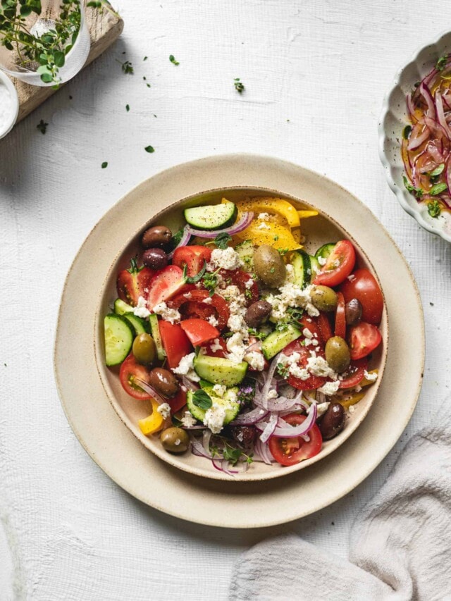 A colorful Greek salad on a white plate, featuring tomatoes, cucumbers, olives, red onions, bell peppers, crumbled feta cheese, and a sprinkle of herbs. The salad is set on a textured white tabletop with additional ingredients nearby.