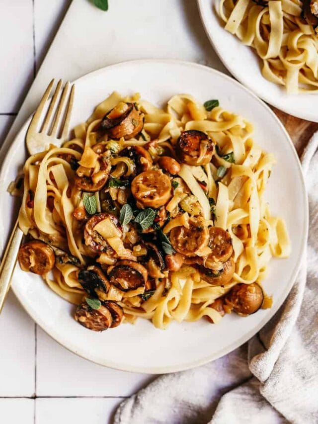 A plate of tagliatelle pasta topped with browned sausage slices and fresh herbs. A fork rests on the plates side. The dish is set on a white tiled surface with part of a beige cloth napkin visible.