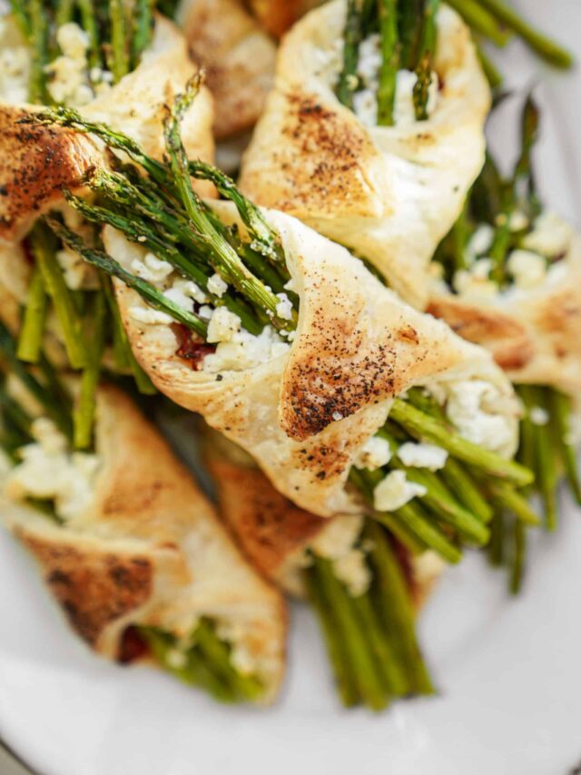 Close-up of puff pastry bundles filled with asparagus and crumbled cheese. The pastries are golden brown with a speckled seasoning, arranged on a white plate.