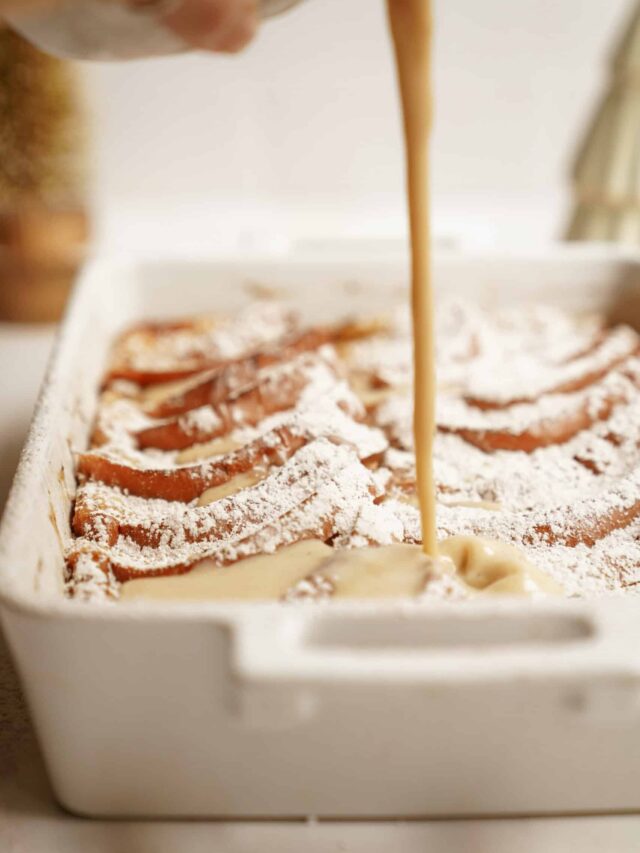 A hand pours creamy sauce over a white casserole dish filled with baked French toast, dusted with powdered sugar. The dish sits on a kitchen counter, suggesting a warm, homemade breakfast setting.