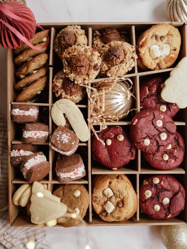 A box of assorted cookies and treats, including red velvet cookies with white chocolate chips, marshmallow cookies, chocolate-covered marshmallows, tree-shaped sugar cookies, and decorative ornaments.