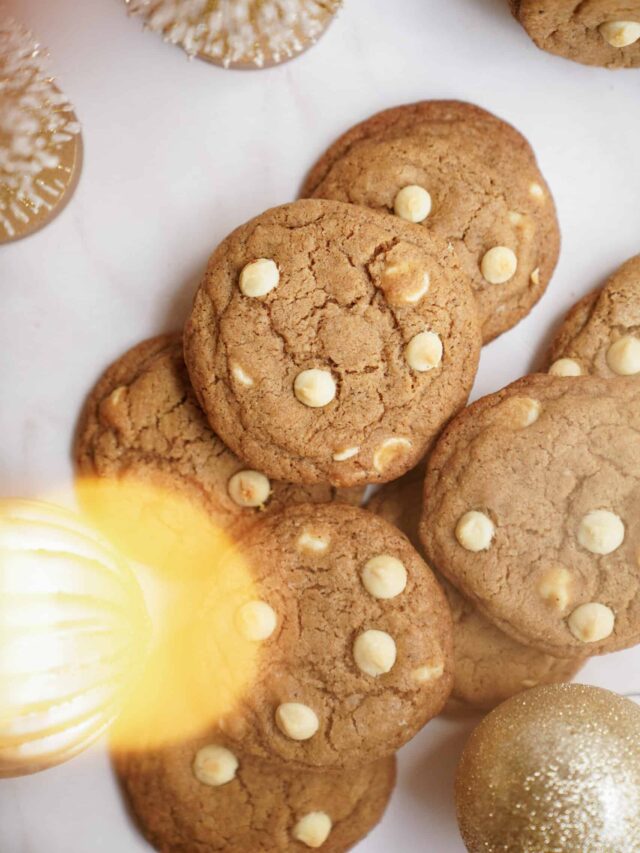 A pile of brown cookies with white chocolate chips on a light surface. The cookies are surrounded by decorative elements, including gold ornaments and small white Christmas trees.