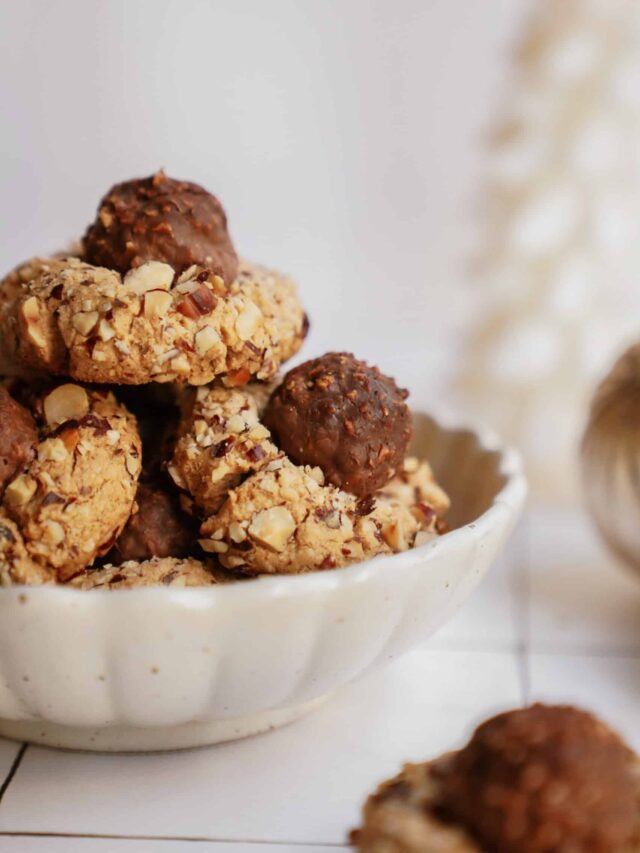 A white bowl filled with cookies coated in chopped nuts and topped with chocolate truffles. The cookies are stacked, highlighting the textured surface and rich colors. Subtle background with soft lighting.