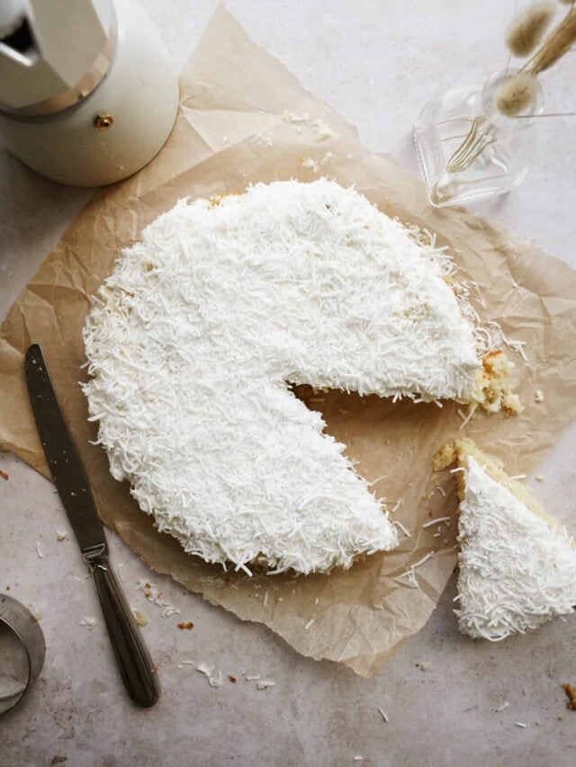 A round coconut cake with a single slice removed, displayed on parchment paper. A knife and a piece of cake are beside it. The background includes a white teapot and a small glass vase with dried flowers.