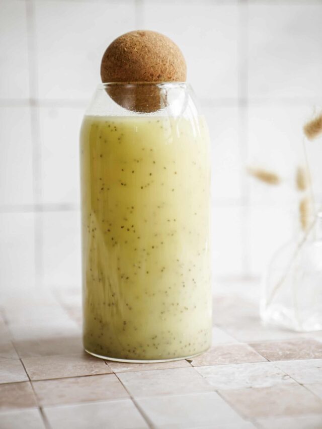 A glass bottle filled with a light green chia seed beverage, sealed with a cork stopper, stands on a tiled surface. The background shows a blurred white grid pattern and a small vase with dried wheat.