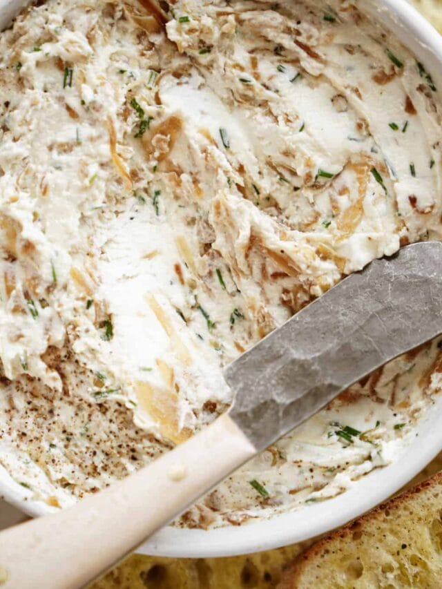 A close-up of a white bowl filled with creamy dip mixed with herbs and caramelized onions. A butter knife with a wooden handle rests on the edge of the bowl. The texture is smooth and speckled with green herbs.