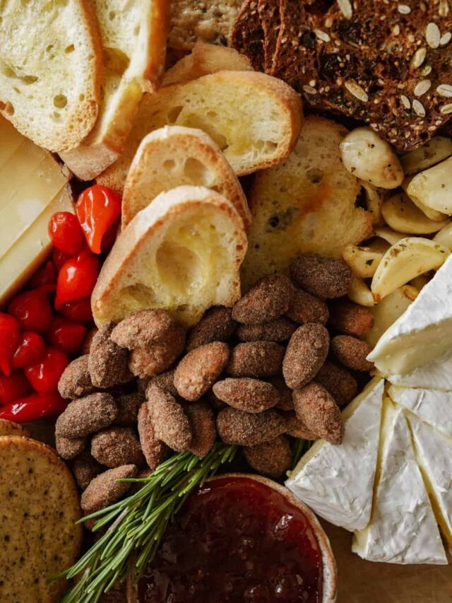 A close-up of a charcuterie board featuring sliced bread, roasted garlic, seasoned almonds, brie cheese, cherry peppers, seeded crackers, and a dish of jam, garnished with sprigs of rosemary.