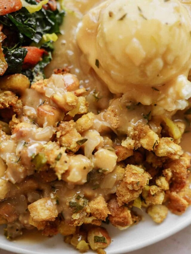 A plate of Thanksgiving-style food featuring mashed potatoes with gravy, stuffing, and a side of sautéed leafy greens with diced carrots. The meal is richly garnished and set on a white background.