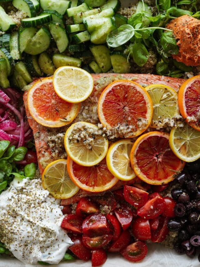 A colorful platter with a baked trout filet topped with lemon and orange slices, surrounded by fresh vegetables