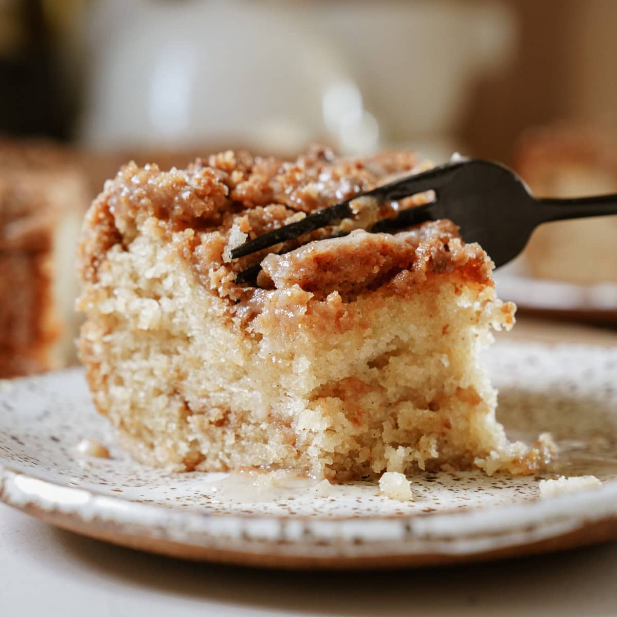 A slice of golden-brown coffee cake on a white plate, with a black fork cutting through it. 