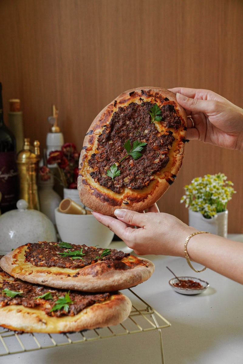 Hands holding up a round Manakeesh flatbread, showcasing its crispy edges and generous topping of seasoned ground beef. Fresh herbs are sprinkled on top, adding pops of green against the golden-brown bread and savory meat.