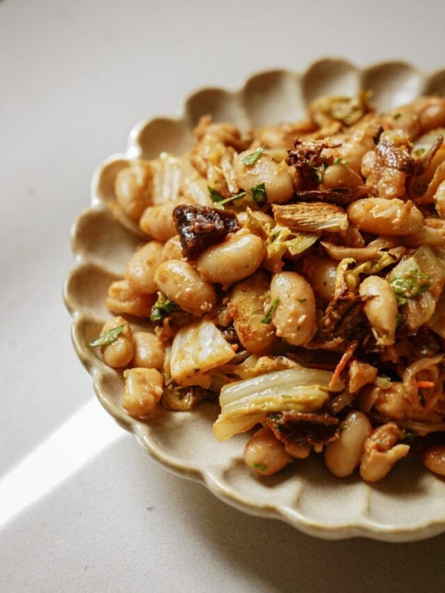 A plate of seasoned white beans with chopped vegetables and herbs, served on a scalloped-edged dish against a neutral background. The dish includes visible bits of sautéed onions and mushrooms.