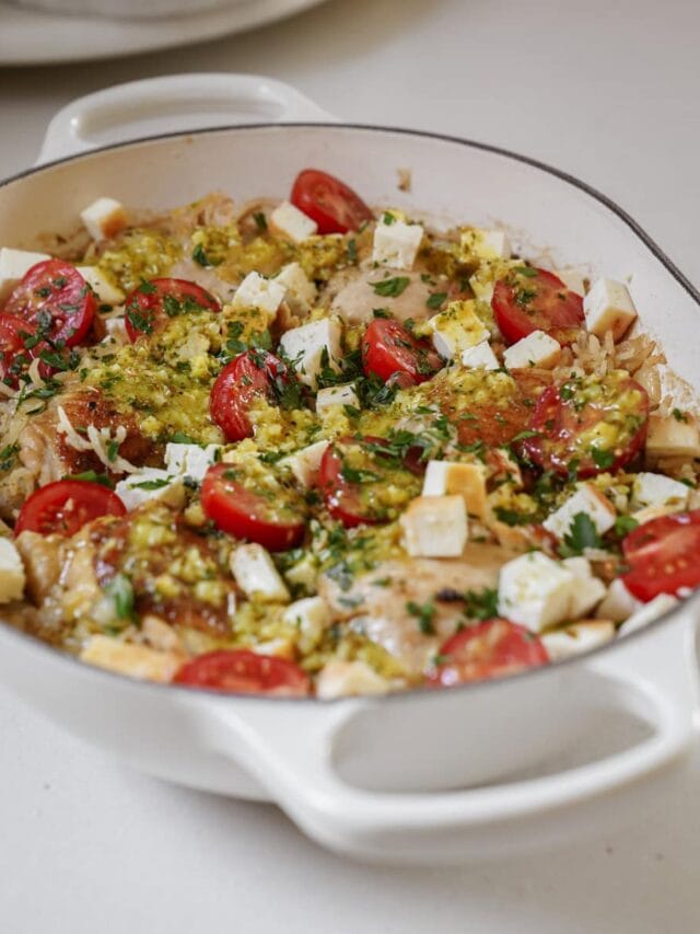 A white casserole dish filled with baked chicken, topped with cherry tomatoes, diced cheese, herbs, and a green sauce. The dish is placed on a light-colored surface.