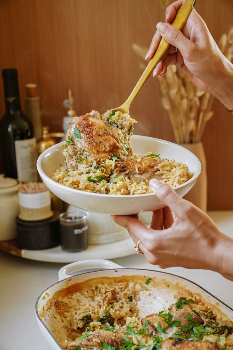 Hands cradle a white bowl filled with a golden, creamy Cheese Chicken and Rice Casserole. The dish showcases tender pieces of chicken nestled among cheesy rice, with flecks of herbs visible throughout.