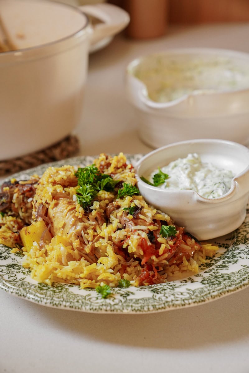 A white and blue plate piled high with golden Chicken Biryani and a bowl of tzatziki sauce.