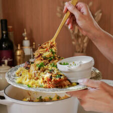 A hand holds a gold spoon above a white and blue plate piled high with golden Chicken Biryani and a bowl of tzatziki sauce.