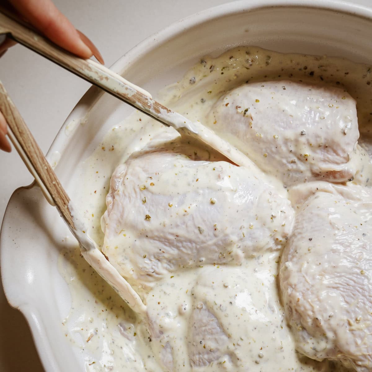 A white ceramic baking dish contains raw chicken pieces coated in a creamy, herb-flecked Greek yogurt marinade. 