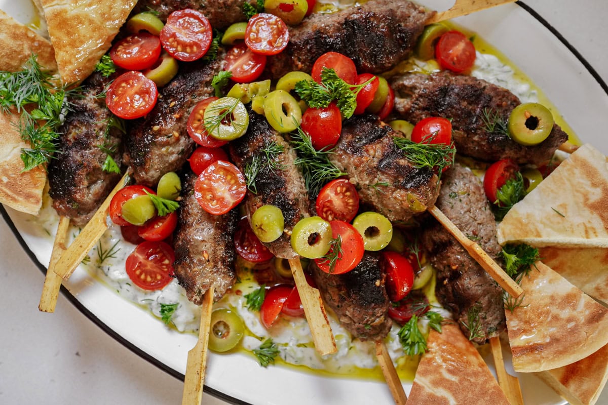 A platter of grilled beef kofta kebabs and pita on tzatziki, topped with tomatoes and olives.