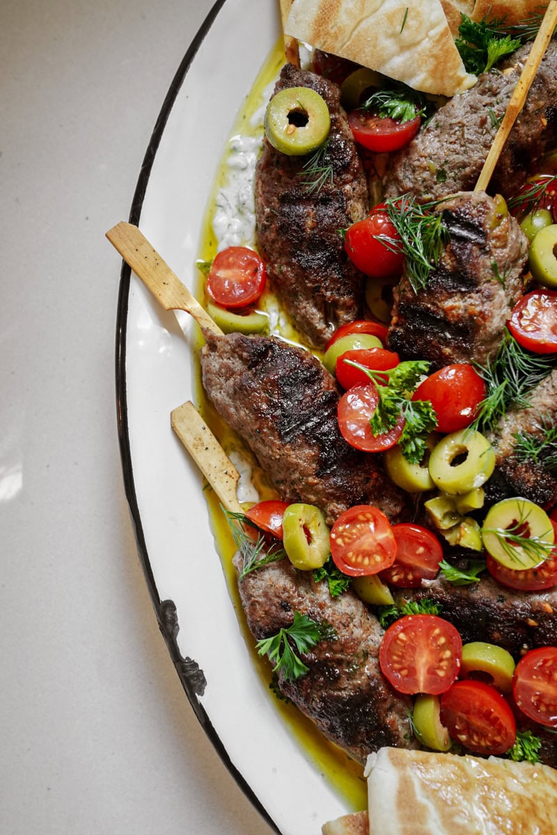 The side of a platter of grilled beef kofta kebabs on tzatziki, topped with tomatoes and olives.