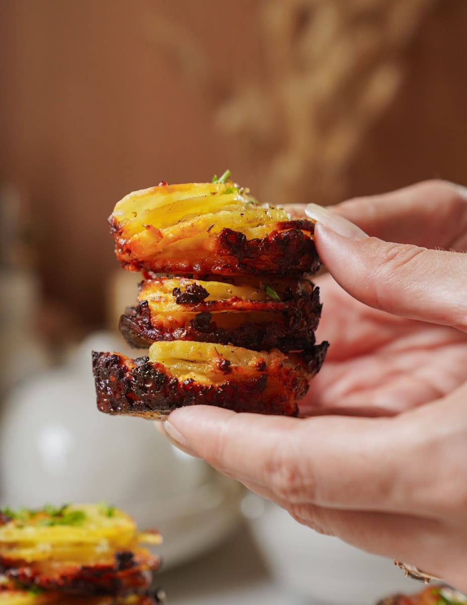 A person holds a stack of crispy potato slices, seasoned and golden brown, garnished with freshly chopped herbs. The background is softly blurred, highlighting the focus on the savory snack.