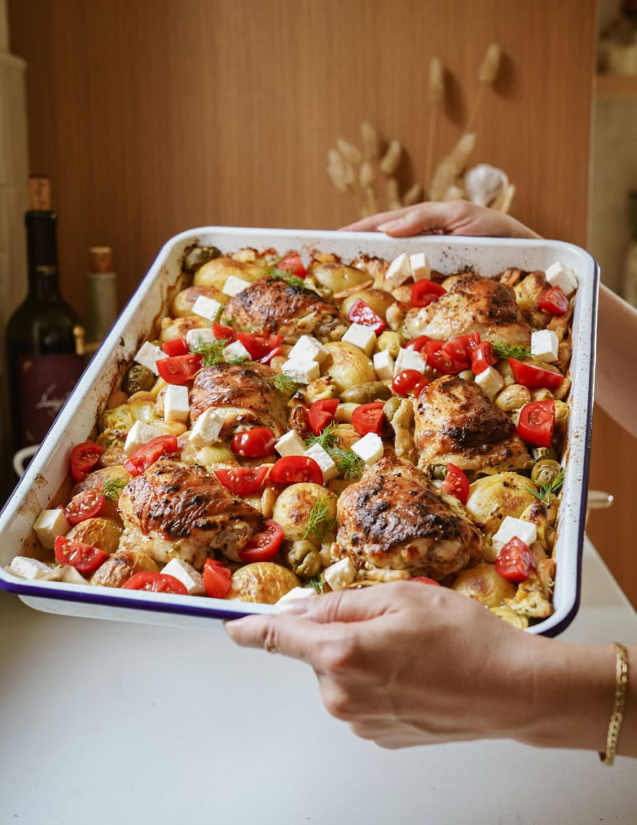 Hands hold a sheet pan displaying a colorful array of Greek-inspired ingredients: crispy-skinned roasted chicken thighs nestled among golden-brown potato wedges, green olives, and tender artichoke hearts. The dish is garnished with vibrant red cherry tomato halves and scattered cubes of white feta cheese.