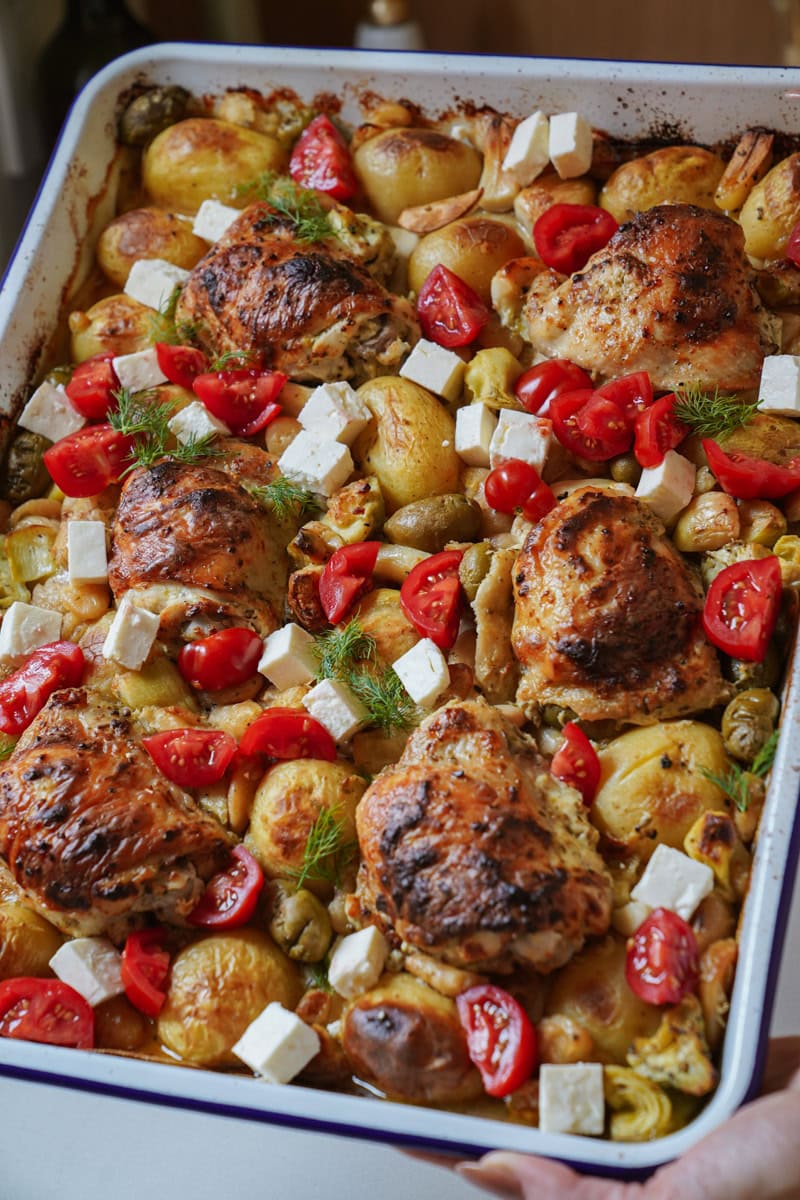 A sheet pan displays a colorful array of Greek-inspired ingredients: crispy-skinned roasted chicken thighs nestled among golden-brown potatos, green olives, and tender artichoke hearts. The dish is garnished with vibrant red cherry tomato halves and scattered cubes of white feta cheese.