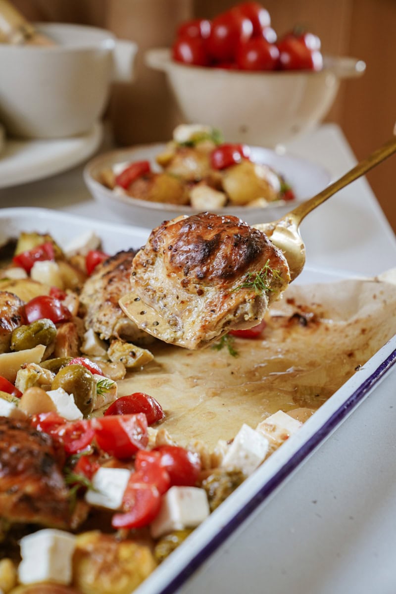 A gold spoon lifts a perfectly roasted chicken thigh above a sheet pan filled with colorful Mediterranean ingredients. The chicken's skin is golden-brown and crispy, glistening with herbs and spices. Below, the pan showcases a medley of roasted potatoes, olives, artichokes, and butter beans, garnished with bright cherry tomatoes and feta.