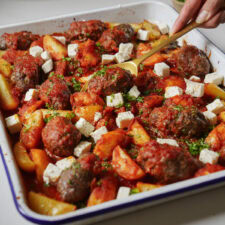 A baking dish of Soutzoukakia filled with roasted meatballs, chunks of potatoes, and diced white cheese, all garnished with fresh herbs. A hand holds a spoon, poised to serve the dish. The ingredients are generously covered in a rich red sauce.