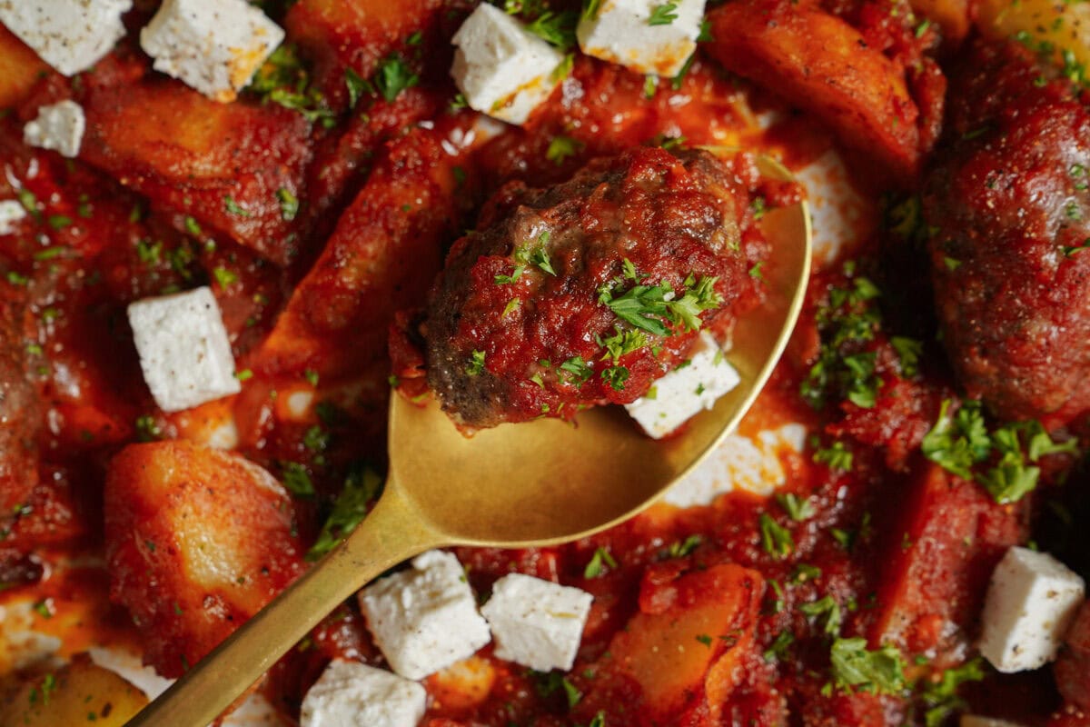 Close-up of a dish featuring browned meatballs in rich tomato sauce, accompanied by cubed feta cheese and garnished with chopped parsley. A golden spoon rests in the center, surrounded by chunks of potato.