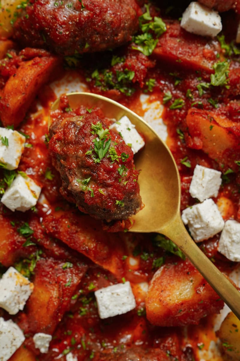 Close-up of a dish featuring browned meatballs in rich tomato sauce, accompanied by cubed feta cheese and garnished with chopped parsley. A golden spoon rests in the center, surrounded by chunks of potato.