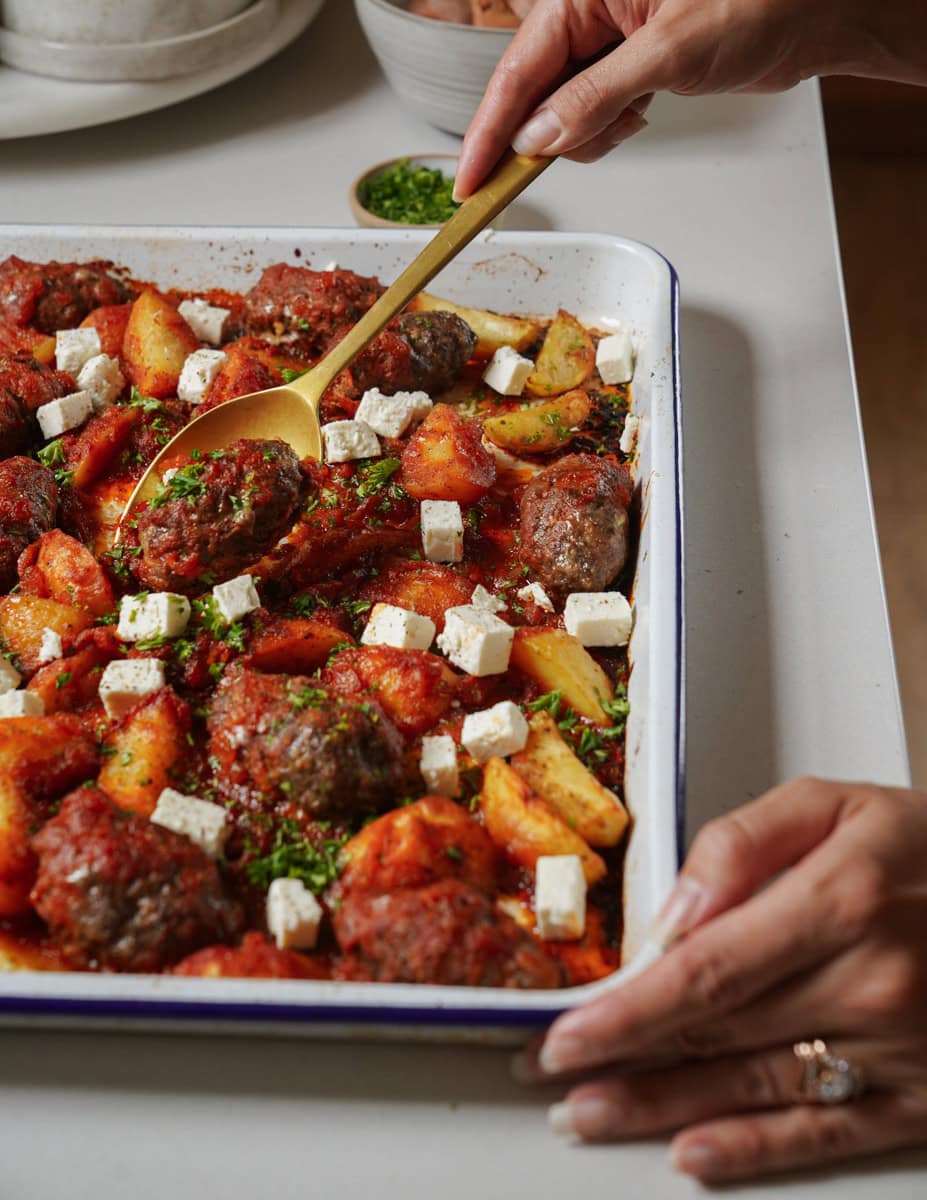 A hand holding a spoon serves baked meatballs, potatoes, and tomato sauce from a baking dish. The dish is topped with white cheese cubes and sprinkled with green herbs. The scene is set on a light-colored countertop.