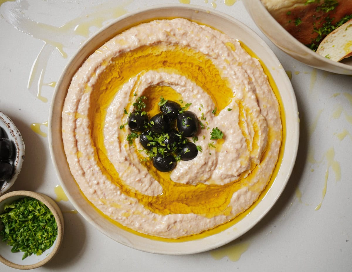 A bowl of creamy hummus garnished with black olives and chopped parsley. Olive oil is drizzled on top, and there are small bowls of chopped parsley and olives nearby on a light surface.