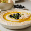 A bowl of creamy hummus garnished with black olives and fresh herbs, drizzled with olive oil. The dish is set on a neutral-colored surface, with a small bowl of black olives in the background.