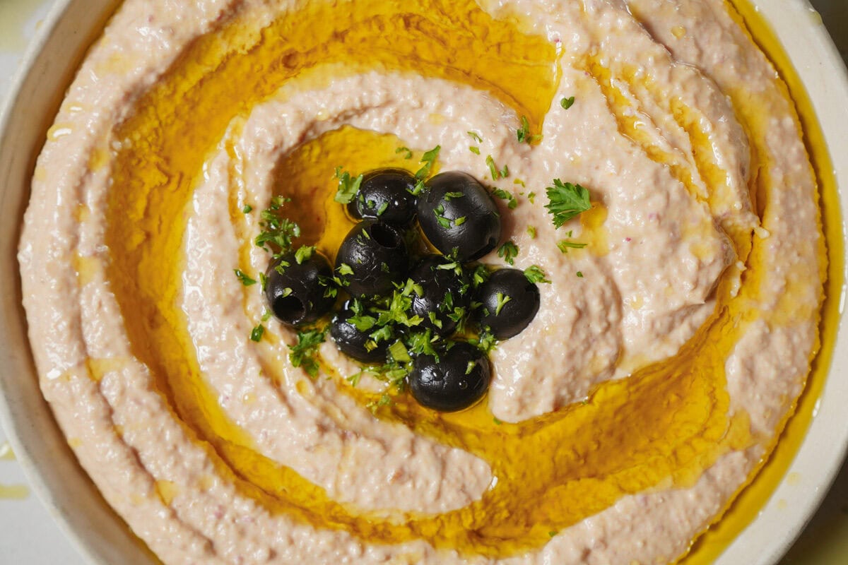 A close-up of a bowl of creamy hummus swirled with olive oil, topped with a small pile of black olives and sprinkled with chopped parsley. The dish has a smooth texture and a warm, inviting appearance.