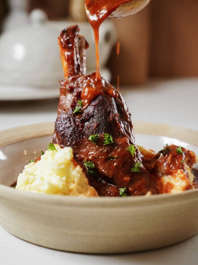 A succulent lamb shank drizzled with rich sauce sits atop creamy mashed potatoes in a beige bowl. Sprinkles of fresh green herbs garnish the dish, with a teapot blurred in the background.