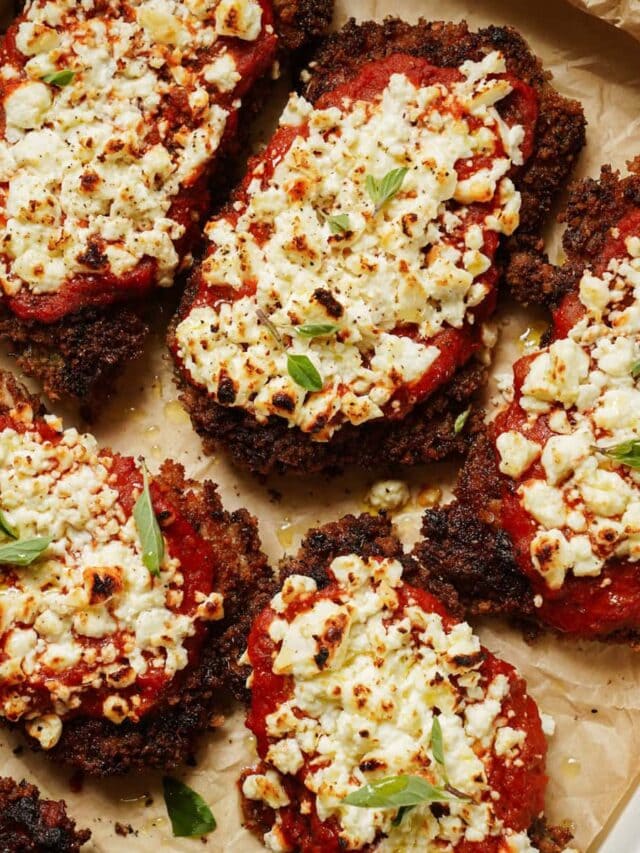 Close-up of crispy breaded chicken cutlets topped with tomato sauce and crumbled feta cheese, garnished with fresh herbs on a parchment-lined tray. The dish is golden brown with hints of green from the herbs.